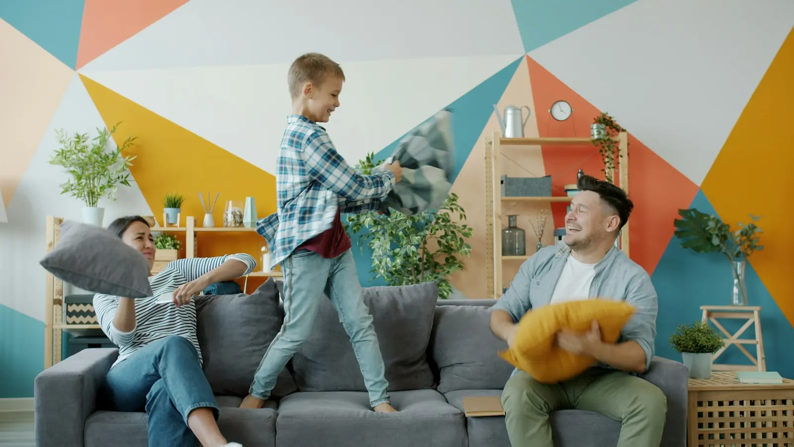 A boy standing on the sofa in a pillow fight with his mum and dad, in front of a brightly painted wall
